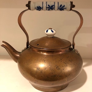 May include: A vintage copper teapot with a white and blue floral patterned handle and lid knob. The teapot has a spout and a curved handle. The copper has a warm, aged patina, suggesting it's an antique or vintage piece.