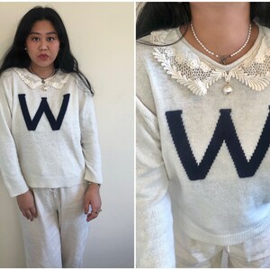 May include: White sweater with a large blue "W" on the front. The sweater has a white lace collar and a pearl necklace.