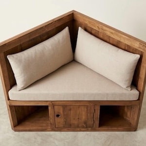 May include: A corner bench constructed from medium-toned wood featuring a triangular seat cushion and two rectangular back cushions in a light beige fabric. The base includes two open storage compartments flanking a central cabinet door with a circular pull.