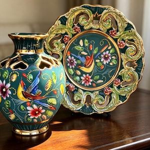 May include: A dark teal ceramic vase and a matching decorative plate, both featuring gold-trimmed edges, intricate cut-out details, and hand-painted motifs of colourful birds, green leaves, and small flowers. The text Dola Al appears in the corner.