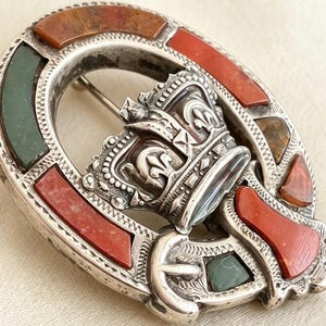 May include: A silver-toned brooch shaped like a buckle, featuring a central crown emblem. The frame is inlaid with alternating segments of smooth, polished red and dark green stones, set within a detailed, textured metal border.