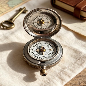 May include: A round, silver-toned metal pocket compass with a hinged lid that is open, revealing a white dial with black cardinal directions including N, E, S, and W. The compass features a central needle and a small, rounded metal knob at the base.