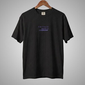 May include: A black short-sleeve t-shirt featuring the text "Emilique sport" in purple lettering on the center chest, accompanied by a small purple checkered pattern graphic positioned directly underneath the text.