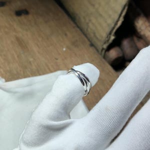 Può includere: Un anello in argento lucido caratterizzato da un design a tre fasce, in cui tre bande lisce e arrotondate sono intrecciate per formare un unico pezzo coeso. Il metallo presenta una finitura brillante e riflettente che cattura la luce.