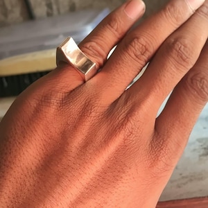 May include: A polished silver-toned ring with a thick, angular, and geometric band design. The ring features a flat, rectangular top surface that sits prominently on the finger, showcasing a smooth, reflective metallic finish throughout.