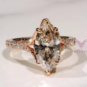 Peut inclure: Une bague en or rose avec une grande pierre centrale taille marquise maintenue par des griffes, ornée de petites pierres rondes sur l'anneau. Le bijou présente le texte VELORA avec un logo en forme de V rose sur le côté droit.