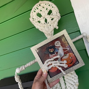 May include: A macrame skeleton decoration with a framed Halloween picture. The skeleton is made of white rope and features a skull-shaped head. The framed picture has a silver glitter frame and depicts a skeleton, an owl, and pumpkins.