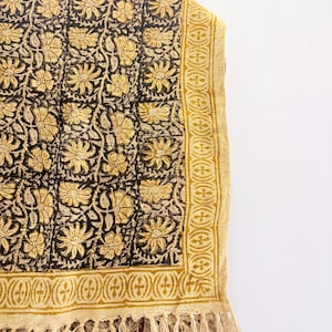 May include: A rectangular textile featuring a dark brown background with a repeating golden yellow floral pattern. The fabric has a wide golden yellow border with a geometric design and a fringe edge along the bottom, showcasing a woven texture.