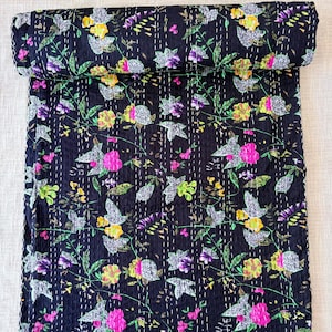 May include: A black quilt featuring a floral pattern with yellow, pink, and purple flowers, green leaves, and white accents. The fabric is decorated with rows of white running stitches that create a textured, quilted appearance throughout.