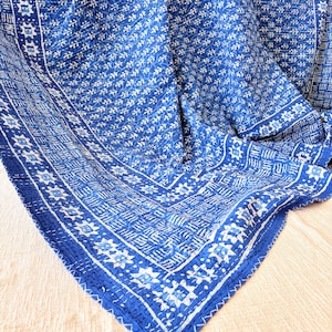 May include: A blue and white textile featuring a repeating floral pattern across the center and a geometric border design. The fabric displays visible white stitching throughout, creating a quilted texture with a layered, folded appearance.