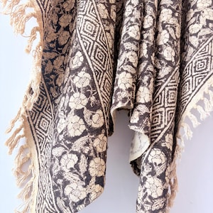 May include: A textile featuring a dark brown and beige floral pattern with a geometric diamond border. The fabric has a textured appearance and is finished with long, knotted fringe along the edges, creating a decorative drape effect.