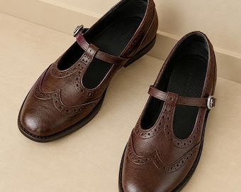 May include: A pair of dark brown T-strap shoes featuring classic wingtip brogue detailing with perforated patterns along the toe cap and sides. Each shoe includes a thin adjustable strap with a small silver-toned metal buckle closure.
