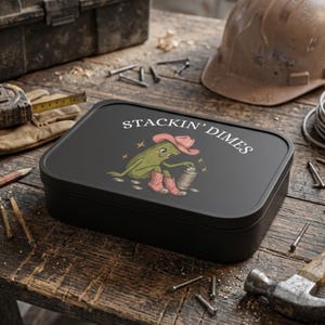 May include: A rectangular black storage tin featuring a graphic of a green pickle wearing a pink cowboy hat and boots, stacking silver coins. The text STACKIN' DIMES is printed in white above the illustration on the lid of the container.