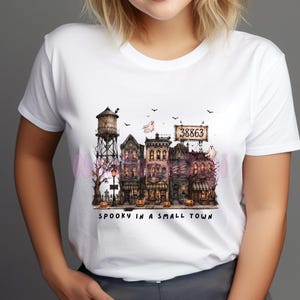 May include: A white short-sleeved t-shirt featuring a graphic print of a spooky town scene with a water tower, old buildings, ghosts, bats, and pumpkins. The text "38863" appears on a sign, and the words "SPOOKY IN A SMALL TOWN" are printed below.