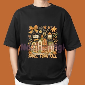 May include: A black t-shirt featuring a graphic print of a small town scene with houses, a water tower, pumpkins, and a bow. The text "260583" appears on a sign, and the words "SMALL TOWN FALL" are printed in orange at the bottom of the design.