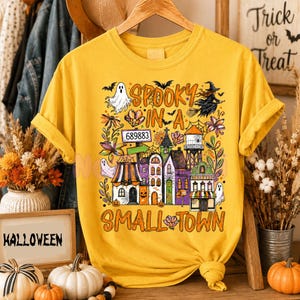 May include: A mustard yellow t-shirt featuring a graphic print of a spooky small town with houses, a water tower, ghosts, bats, and a witch on a broomstick. The text reads SPOOKY IN A SMALL TOWN and includes the number 689883 on a sign.
