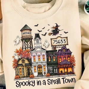 Könnte beinhalten: Ein beigefarbenes Sweatshirt mit einer Grafik einer Kleinstadt, einem Wasserturm mit der Aufschrift Carthage, einer Werbetafel mit der Zahl 75633, Geistern, Fledermäusen, einer Hexe auf einem Besen und Herbstbäumen. Der Text Spooky in a Small Town steht unten.