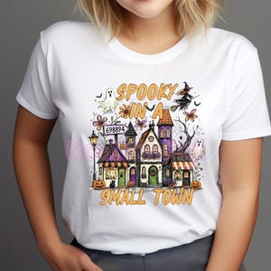 May include: A white t-shirt featuring a graphic print of a small town with purple and orange buildings, pumpkins, ghosts, a witch on a broomstick, and the text "SPOOKY IN A SMALL TOWN" with a sign displaying the number 698894 above the houses.