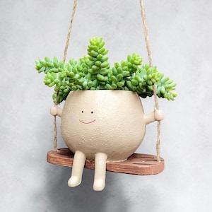 May include: A beige ceramic planter with a textured surface, a simple smiling face, and two legs, sitting on a small wooden swing suspended by twine. The planter holds a vibrant green Sedum morganianum succulent plant with trailing stems.