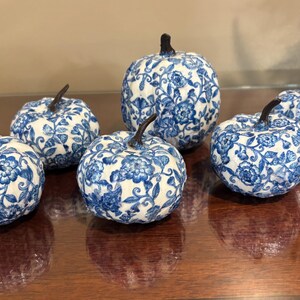 May include: A collection of five decorative pumpkins featuring a white base with intricate blue floral patterns and dark brown stems. The pumpkins vary in size and are arranged in a group, each displaying a detailed botanical design across the surface.
