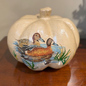 May include: A cream-colored ceramic pumpkin figurine featuring a detailed illustration of two ducks swimming in blue water with green reeds. The pumpkin has a smooth, glossy finish and a textured surface with visible ridges and a central stem.