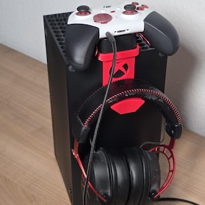 Könnte beinhalten: Ein schwarzer Spielkonsolenständer mit roter Halterung für einen weiß-schwarzen Wireless-Controller und einem roten Haken für schwarz-rote Over-Ear-Kopfhörer. Die Konsole ist rechteckig mit einem Gittermuster oben und einem roten Logo.