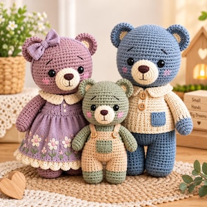 Könnte beinhalten: Drei gehäkelte Teddybären in verschiedenen Größen. Der linke Bär ist lila mit einem Blumenkleid und einer Schleife. Der mittlere Bär ist grün und trägt eine beige Latzhose. Der rechte Bär ist blau und trägt ein cremefarbenes Hemd mit Knöpfen.