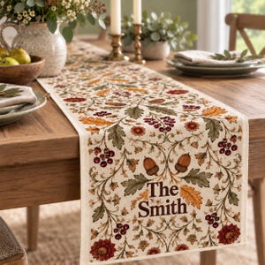 May include: A cream-colored table runner featuring a repeating botanical pattern of brown acorns, green oak leaves, and red berries. The text "The Smith" is printed in a dark brown serif font near the bottom edge of the fabric runner.