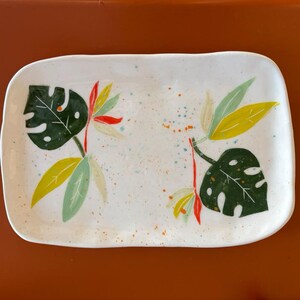 Puede incluir: Una bandeja rectangular de cerámica con esquinas redondeadas, que presenta una base blanca con detalles de pintura salpicada. La superficie está decorada con ilustraciones pintadas de hojas de monstera verde oscuro, hojas amarillas y acentos rojos.