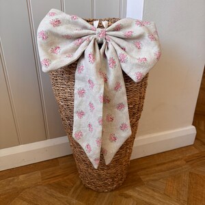 May include: A large decorative bow made of beige fabric with a repeating pink floral pattern. The bow features two wide loops and two long, pointed tails hanging downward, secured at the centre with a matching fabric knot, resting on a woven basket.