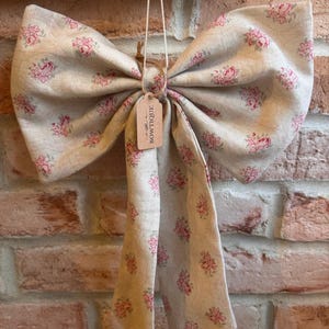 May include: A large decorative bow made of beige fabric with a repeating pink floral pattern. The bow features two long hanging tails and a small rectangular cardboard tag attached by a string, which has the word BOWTIQUE printed on it.