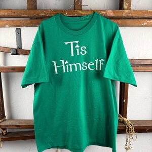 Può includere: Una t-shirt verde brillante a maniche corte con la scritta bianca 'Tis Himself' stampata sul petto. La lettera 'i' di 'Tis' è sostituita da un piccolo grafico a forma di trifoglio bianco. La maglia ha un girocollo e un orlo dritto.