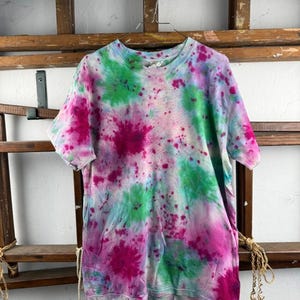 Puede incluir: Una camiseta de manga corta con un patrón de teñido anudado que presenta salpicaduras vibrantes de color rosa intenso, verde brillante y azul claro por toda la tela. La prenda tiene cuello redondo y se muestra en una percha de alambre.