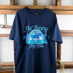 Può includere: Una t-shirt blu navy a maniche corte con una stampa grafica recante il testo Ha' Penny Horse Trials April 8-10, 1994. Il design include una silhouette stilizzata di un cavallo e un cavaliere nei toni del blu, del verde acqua e del bianco.
