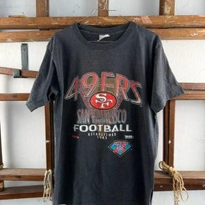 Può includere: Una t-shirt grigio scuro a maniche corte con una stampa grafica che recita 49ERS SAN FRANCISCO FOOTBALL ESTABLISHED 1945, con un logo della squadra rosso e oro al centro e un piccolo emblema a forma di diamante blu e rosso vicino all'orlo.