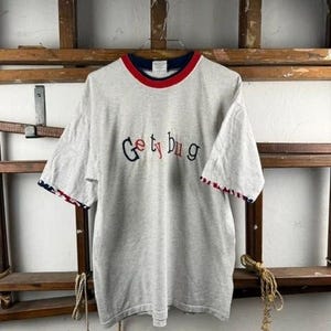 Può includere: Una t-shirt a maniche corte grigio chiaro con il testo ricamato "Ge ty bug" in lettere nere e rosse. La maglia presenta un colletto a strati blu navy e rosso, e i bordi delle maniche hanno una finitura con motivo rosso, bianco e blu.