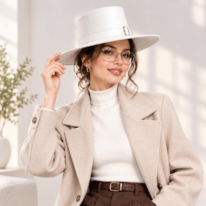 May include: A white wide-brimmed hat with a flat crown and a matching band featuring a silver-toned metal buckle. The hat is worn with a white ribbed turtleneck sweater, a beige wool-blend coat, and brown trousers with a dark brown leather belt.