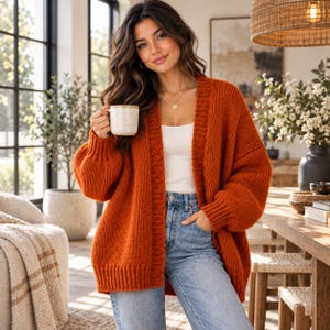 Peut inclure: Un gilet ouvert en tricot épais orange brûlé avec des manches longues et des poignets côtelés, porté sur un débardeur blanc et un jean bleu clair. Le gilet présente un motif en tricot texturé épais et une coupe décontractée et ample.