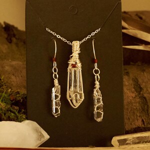 Op de afbeelding: Een sieradenset bestaande uit een ketting met hanger en twee bijpassende oorbellen. Elk stuk bevat een helder, ruw kristalpunt gewikkeld in zilverdraad met een kleine rode kraal als accent, hangend aan een fijne ketting van zilverkleurig metaal.