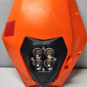 May include: A bright orange motorcycle headlight mask featuring a central black housing that contains four circular LED lights arranged in a square pattern. The plastic orange shroud has a smooth finish with a small red square decal on the left side.