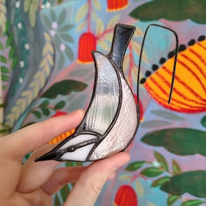 May include: A stained glass ornament shaped like a bird, featuring iridescent white and textured clear glass panels held together by dark metal solder lines, with a curved metal wire hook attached to the top for hanging purposes.