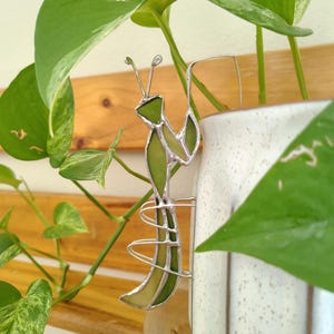 Peut inclure: Un ornement en vitrail représentant une mante religieuse, composé de segments de verre vert clair assemblés par des lignes de soudure métallique argentée. La figurine possède deux antennes et des pattes courbées pour s'accrocher.