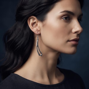May include: A silver-toned metal drop earring featuring a curved, elongated shape with a hollow center. The earring hangs from a simple hook wire, showcasing a smooth, polished finish that catches the light, creating a sleek and modern appearance.