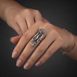 May include: A silver ring featuring an elongated, S-shaped design with two fan-shaped, textured ends. The metal has a polished finish with darkened recessed areas to highlight the intricate, radiating line details on the fan-shaped sections.