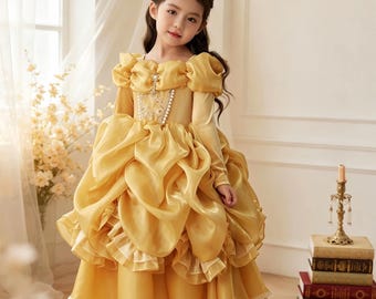 May include: A golden yellow ball gown featuring off-the-shoulder puffed sleeves, a bodice with vertical pearl-like trim, and a voluminous skirt with layered ruffles and lace hem detailing. A matching golden yellow headpiece is worn.