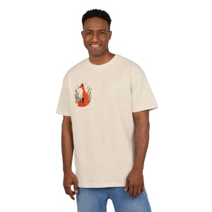 以下が含まれることがあります： 左胸に小さなグラフィックが施されたクリーム色の半袖Tシャツ。グラフィックには、濃い緑色のシダのような葉と小さなオレンジ色のドットの中に座っている、様式化されたオレンジ色のキツネが描かれています。