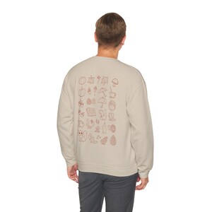 Könnte beinhalten: Ein hellbeiges Sweatshirt mit Rundhalsausschnitt und einem Raster aus kleinen, rotbraunen Strichzeichnungen auf der Rückseite. Die Grafiken zeigen verschiedene herbstliche Symbole wie Kürbisse, Blätter, Pilze, Eicheln, ein Eichhörnchen und eine Kaffeetasse.