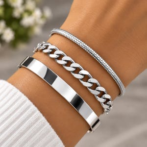 May include: A set of three silver-toned bracelets worn together. The set includes a smooth, flat cuff, a medium-sized curb chain bracelet, and a thin, textured snake chain bracelet, all featuring a polished metallic finish.