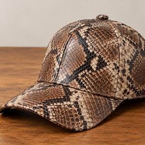 May include: A baseball cap featuring a brown, black, and cream snakeskin pattern. The cap has a structured six-panel crown with a matching button at the top and a curved brim, showcasing a detailed reptilian scale texture across the entire surface.