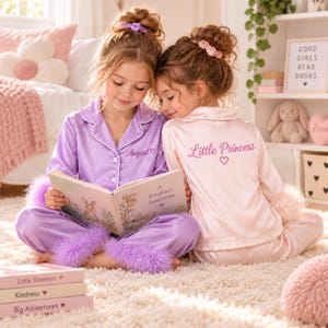 May include: Two children wearing matching satin pajama sets with feather trim on the cuffs and ankles. One set is lavender with the name August and a heart, the other is pale pink with the text Little Princess and a heart.